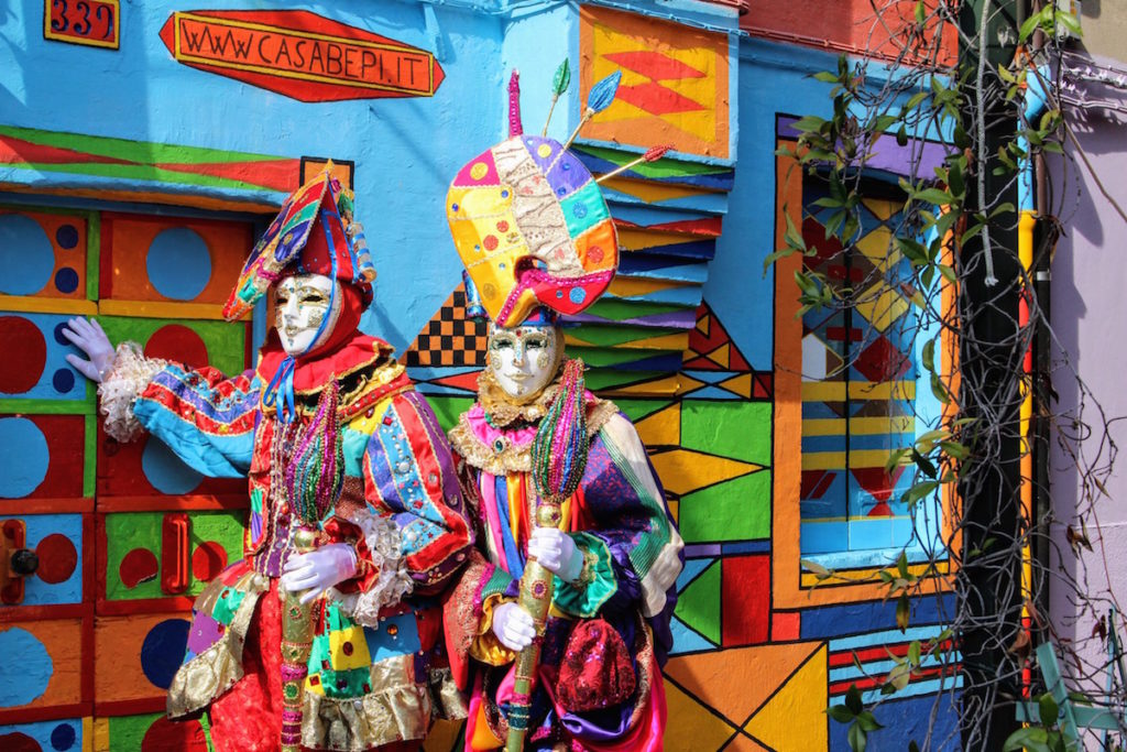 A very colorful couple dressed in traditional Venetian costumes in Burano, Italy outside the most colorful house on the island