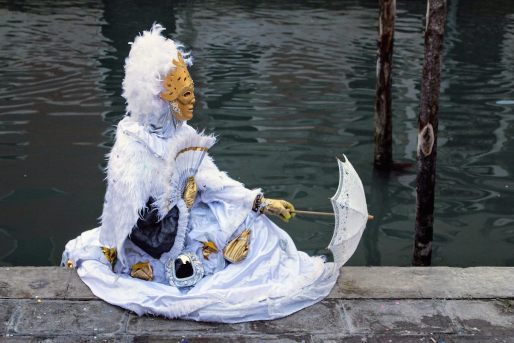 A woman dressed in a traditional white costume with an umbrella sits by a canal in Venice during Carnival