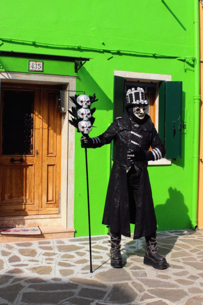 A man with a skull cane and large top hat poses in his traditional costume in front of a green house