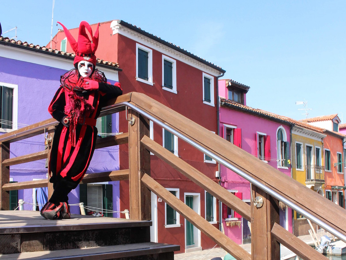 A person in a red Joker costume poses on a bridge in Burano, Italy